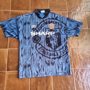 Puede incluir: Una camiseta de f&uacute;tbol azul y negra con el logotipo del Manchester United y la palabra "SHARP" impresa en la parte delantera.