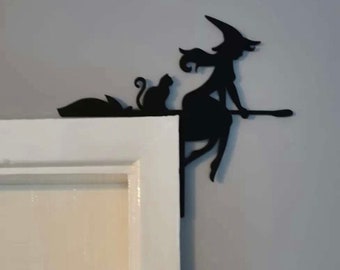 May include: Black silhouette of a witch riding a broomstick with a cat on the broom handle. The witch is wearing a pointed hat and has long flowing hair. The silhouette is mounted on a white door frame.
