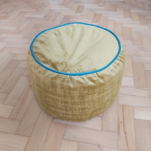 Puede incluir: Un puf redondo de tela amarilla con ribete azul. El puf tiene una superficie texturizada y está sobre un suelo de madera.