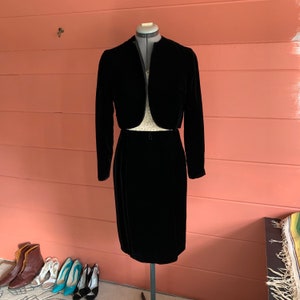 May include: A black velvet skirt suit with a cropped jacket. The jacket has a V-neck and long sleeves. The skirt is knee-length and has a straight silhouette.