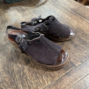 May include: A pair of brown suede sandals with open toes and slingback straps. The sandals have wooden soles and small buckles. The heels are approximately 2 inches high.