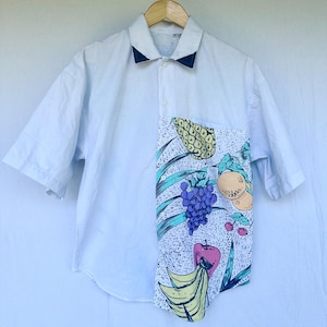 May include: White short-sleeved shirt with a navy blue collar and a pocket featuring a colorful fruit pattern. The fruit pattern includes a pineapple, grapes, bananas, apples, and oranges. The shirt is hanging on a wooden hanger.