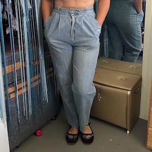 May include: High-waisted light blue denim pants featuring a textured, crinkled fabric finish, a front zipper closure, double belt loops at the waist, and deep side pockets. The pants have a relaxed fit through the hips and legs with a tapered hem.