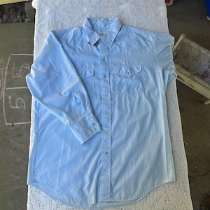 May include: A light blue button-down shirt with long sleeves, two chest pockets, and decorative stitching. The shirt has a classic Western style with snap closures and a pointed collar. The shirt is made of a lightweight fabric.
