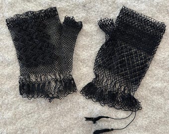 May include: A pair of black fingerless gloves made from a fine, open-weave mesh fabric. The gloves feature a delicate, intricate pattern throughout the material, with a scalloped edge at the wrist and two thin, dangling black thread ties.