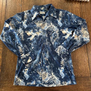 May include: A long-sleeved button-down shirt with a marbled pattern in shades of blue, white, and tan. The shirt has a classic collar, buttoned cuffs, and a chest pocket. The fabric appears to be a soft, flowing material.