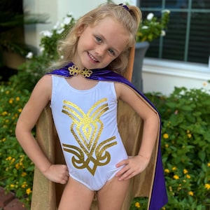 K&ouml;nnte beinhalten: Ein wei&szlig;er Trikotanzug mit goldenem Glitzerdesign und einem goldenen Umhang mit lila Besatz. Das Outfit ist mit einem goldenen Kragen und einem lila Haaraccessoire ausgestattet. Das Outfit ist f&uuml;r Kinder konzipiert.