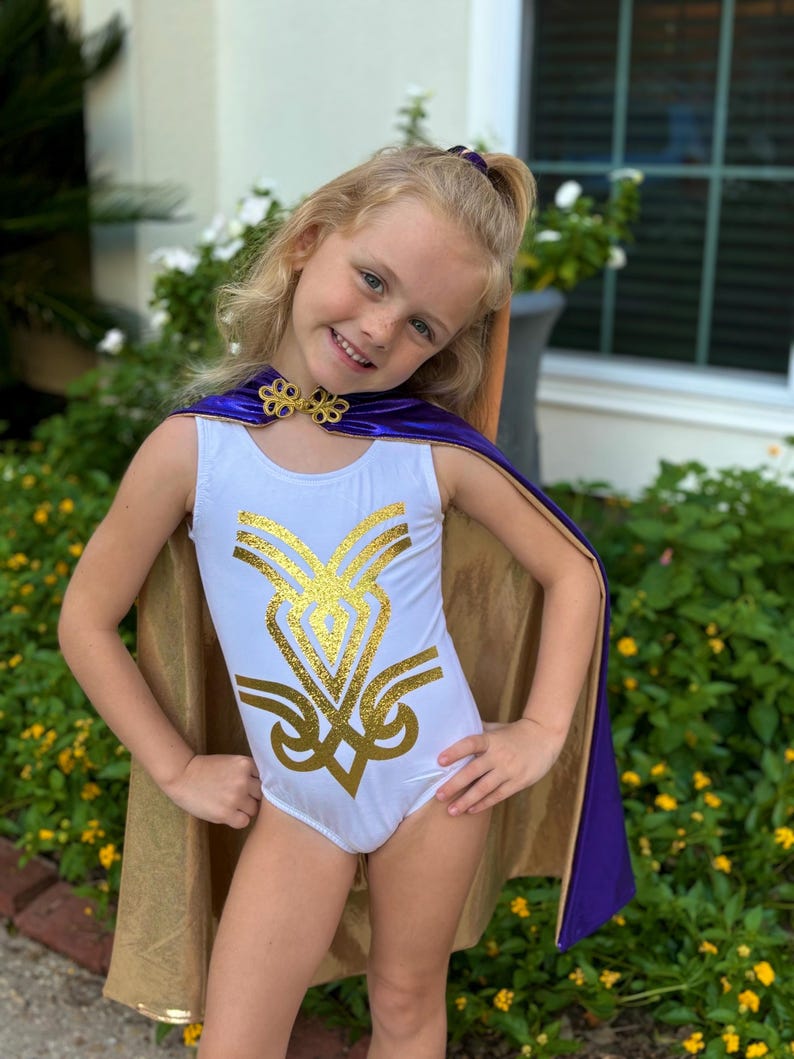 K&ouml;nnte beinhalten: Ein wei&szlig;er Trikotanzug mit goldenem Glitzerdesign und einem goldenen Umhang mit lila Besatz. Das Outfit ist mit einem goldenen Kragen und einem lila Haaraccessoire ausgestattet. Das Outfit ist f&uuml;r Kinder konzipiert.