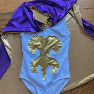 K&ouml;nnte beinhalten: Ein wei&szlig;er Body mit goldenem Emblem und einem lila-goldenen Umhang. Der Umhang hat einen goldenen Verschluss. Der Body ist &auml;rmellos. Der Umhang ist &uuml;ber einer beigefarbenen Oberfl&auml;che drapiert.