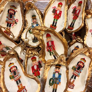 Puede incluir: Adornos navideños hechos con conchas de ostra. Cada concha tiene un borde dorado y presenta un soldado cascanueces pintado. Los adornos están ensartados con hilo dorado y una perla.