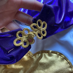 K&ouml;nnte beinhalten: Nahaufnahme eines Kost&uuml;mteils mit gl&auml;nzendem lila Stoff mit goldenen Verzierungen und Akzenten. Die goldene Verzierung hat ein Blumenmuster. Das Kost&uuml;m scheint aus gl&auml;nzendem goldenem Stoff und wei&szlig;em Stoff zu bestehen.