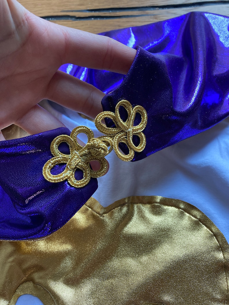 K&ouml;nnte beinhalten: Nahaufnahme eines Kost&uuml;mteils mit gl&auml;nzendem lila Stoff mit goldenen Verzierungen und Akzenten. Die goldene Verzierung hat ein Blumenmuster. Das Kost&uuml;m scheint aus gl&auml;nzendem goldenem Stoff und wei&szlig;em Stoff zu bestehen.