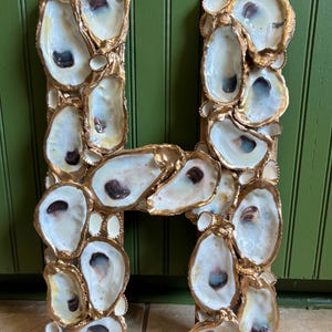 Puede incluir: Una letra "H" decorativa hecha de conchas de ostra. Las conchas están dispuestas para formar la letra, con un borde dorado. Las conchas son blancas y crema con acentos marrón oscuro y azul.