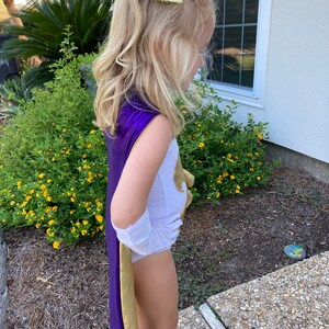 K&ouml;nnte beinhalten: Ein Kind tr&auml;gt einen lila-goldenen Superhelden-Umhang, einen wei&szlig;en Body und wei&szlig;e Handschuhe. Eine gro&szlig;e gelbe Schleife schm&uuml;ckt das blonde Haar des Kindes. Das Kost&uuml;m wird im Freien in der N&auml;he eines Gartens getragen.