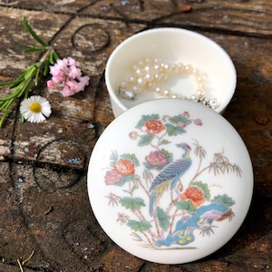 Op de afbeelding: Een witte, ronde sieradendoos met een deksel met een kleurrijk vogel- en bloemmotief. De doos is open en onthult een parelketting. De doos staat op een rustiek houten oppervlak, met bloemen en groen in de buurt.