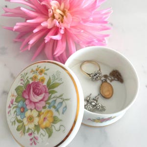 Può includere: Una scatola portaoggetti in porcellana bianca con un design floreale e un bordo dorato. Il coperchio presenta un bouquet di fiori rosa, gialli e blu. La scatola è aperta e contiene gioielli, tra cui un anello d'oro, orecchini e una collana.