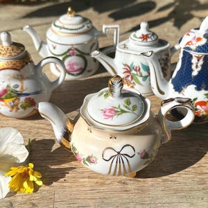 May include: A collection of five miniature porcelain teapots featuring various decorative patterns, including floral motifs, gold accents, and a dark blue scale design. Each teapot has a matching lid and a unique shape, displayed together as a set.
