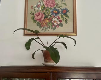 Op de afbeelding: Een ingelijst bloemenborduurwerk met roze, blauwe en witte bloemen hangt boven een donkere houten kast. Een potplant met grote groene bladeren staat bovenop de kast. Het frame is goudkleurig.
