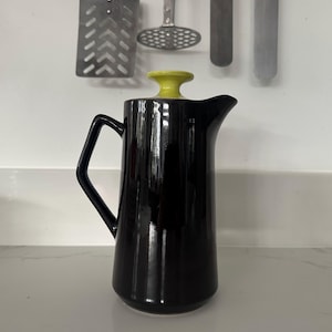 Op de afbeelding: Een glanzende zwarte keramische kan met een gele deksel. De kan heeft een handvat en een tuit. Keukengerei hangt op de achtergrond. De kan wordt waarschijnlijk gebruikt voor het serveren van dranken.