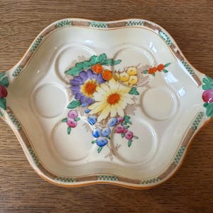 Op de afbeelding: Crèmekleurige keramische eierschaal met zes cirkelvormige inkepingen. Het heeft een bloemenontwerp met gele, paarse en blauwe bloemen. De rand heeft een bruin en groen geometrisch patroon en de handvatten zijn versierd met roze fruit.