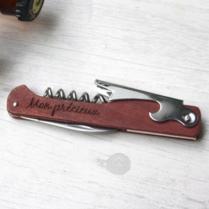 Könnte beinhalten: Ein braunes, mehrteiliges Werkzeug aus Holz mit einem Korkenzieher, einem Flaschenöffner und einem Messer. Das Werkzeug ist mit den Worten "Mon précieux" graviert.