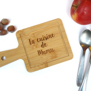 Può includere: Un piccolo tagliere di legno con un manico. Il tagliere ha una superficie di taglio quadrata con un bordo e il testo "La cuisine de Mumu" inciso su di esso.