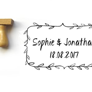 Può includere: Un timbro in legno con una base rettangolare e un manico. Il timbro presenta un bordo decorativo con foglie e il testo "Sophie & Jonathan 18.08.2017".