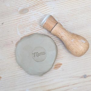 May include: A light brown wooden stamp with a cylindrical handle and a circular base, positioned next to a flat, circular piece of grey clay that features the word Mumu impressed into its center surface in a clear, cursive-style font.
