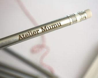 Könnte beinhalten: Ein silberner Bleistift mit einem weißen Radiergummi und der Gravur "Atelier Mumu" auf der Seite.