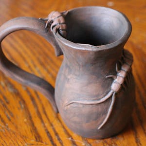 Peut inclure: Une tasse en céramique brune avec une anse et un motif décoratif d'insectes sur le côté.