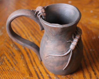 May include: A brown ceramic mug with a handle and a decorative design of insects on the side.