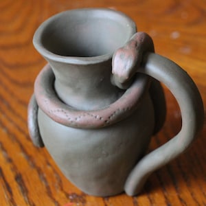 May include: A brown ceramic pitcher with a snake wrapped around the handle and body. The pitcher has a wide mouth and a rounded body.