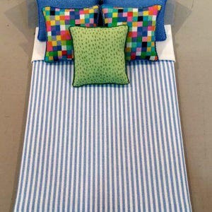 May include: A miniature bed with a blue and white striped comforter. The bed has two blue pillows, two square pillows with a colorful pixelated pattern, and a green pillow with black trim.
