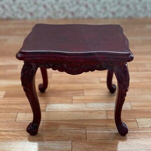 May include: A miniature, dark red wooden table with a square top and ornate, curved legs. The table has a detailed, carved design along the apron and legs, showcasing a classic, antique style. This dollhouse furniture piece is a decorative accent.