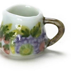 May include: A small white ceramic creamer pitcher featuring a hand-painted fruit motif. The design includes clusters of purple grapes, green leaves, and small red berries encircling the body. It has a single curved handle and a small pouring spout.