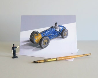 Puede incluir: Un coche de carreras de juguete azul y amarillo con una figura de piloto. El coche está en una tarjeta blanca con un fondo gris.