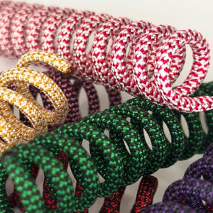 May include: Close-up of coiled rope in various colors, including red and white, gold and white, green and black, and purple and red. The rope has a textured, woven appearance, and the coils are tightly wound. The image is well-lit, highlighting the details of the rope.