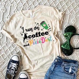 Può includere: Una maglietta beige con il testo "I run on coffee and pixie dust" in lettere nere e colorate. Una fata dei cartoni animati vola sopra il testo. Una tazza di caffè verde con una croce rossa è sopra la fata. La maglietta è appoggiata su una superficie bianca e pelosa con un paio di jeans blu e scarpe da ginnastica bianche e nere.