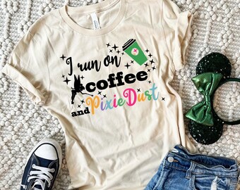 May include: A beige t-shirt with the text "I run on coffee and pixie dust" in black and colorful lettering. A cartoon fairy is flying above the text. A green coffee cup with a red cross is above the fairy. The shirt is laid out on a white fuzzy surface with a pair of blue jeans and black and white sneakers.