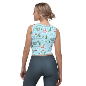 May include: Light blue crop top with a festive pattern featuring castles, palm trees, Christmas trees, and snowflakes. The sleeveless top has a scoop neck and a fitted silhouette. The pattern includes red, green, and white accents, creating a holiday theme.