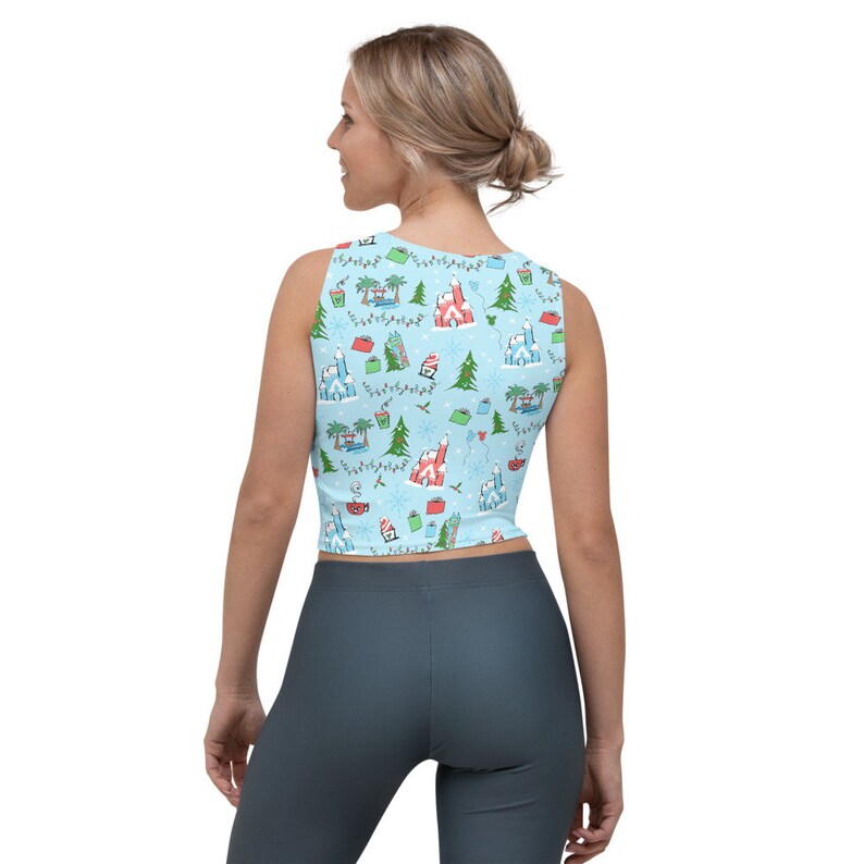 May include: Light blue crop top with a festive pattern featuring castles, palm trees, Christmas trees, and snowflakes. The sleeveless top has a scoop neck and a fitted silhouette. The pattern includes red, green, and white accents, creating a holiday theme.