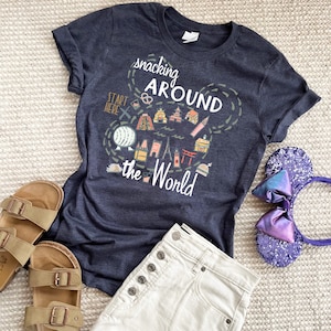 May include: A dark blue t-shirt with a white graphic of a Mickey Mouse head with a map of the world inside. The map includes landmarks from around the world and the text "Snacking Around the World".