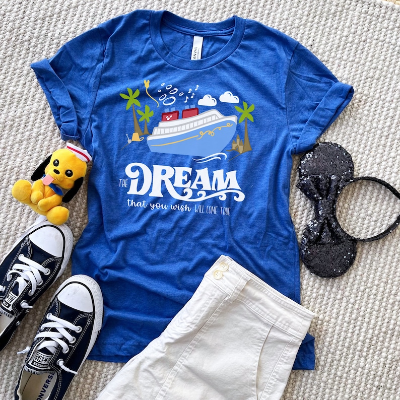 K&ouml;nnte beinhalten: Ein blaues T-Shirt mit einem wei&szlig;en Aufdruck eines Kreuzfahrtschiffs und Palmen. Der Text "The Dream that you wish will come true" ist unter dem Schiff gedruckt.