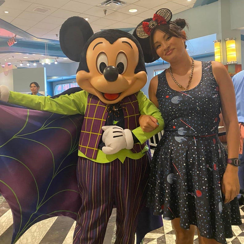 May include: Mickey Mouse dressed in a green and purple costume with a spiderweb cape, posing with a person wearing a black and blue dress with a space-themed pattern.