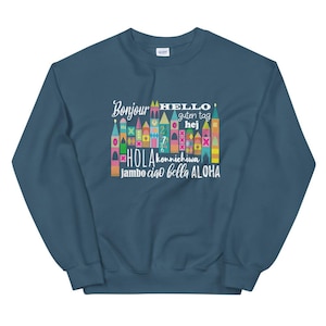 Peut inclure: Un sweat-shirt bleu foncé avec un motif coloré d'un château avec les mots "Bonjour", "Hello", "Guten Tag", "Hej", "Hola", "Konnichiwa", "Jambo", "Ciao", "Bella", et "Aloha" imprimés dessus.