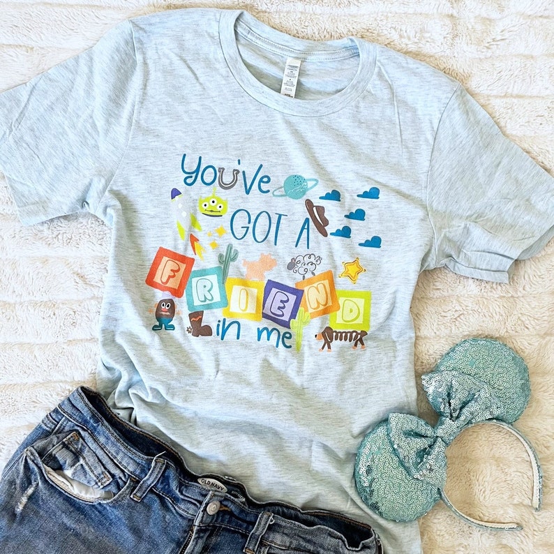 May include: Pale blue t-shirt featuring the text "You've Got a Friend in Me" and cartoon illustrations. The image includes denim shorts and sequined bow ears.