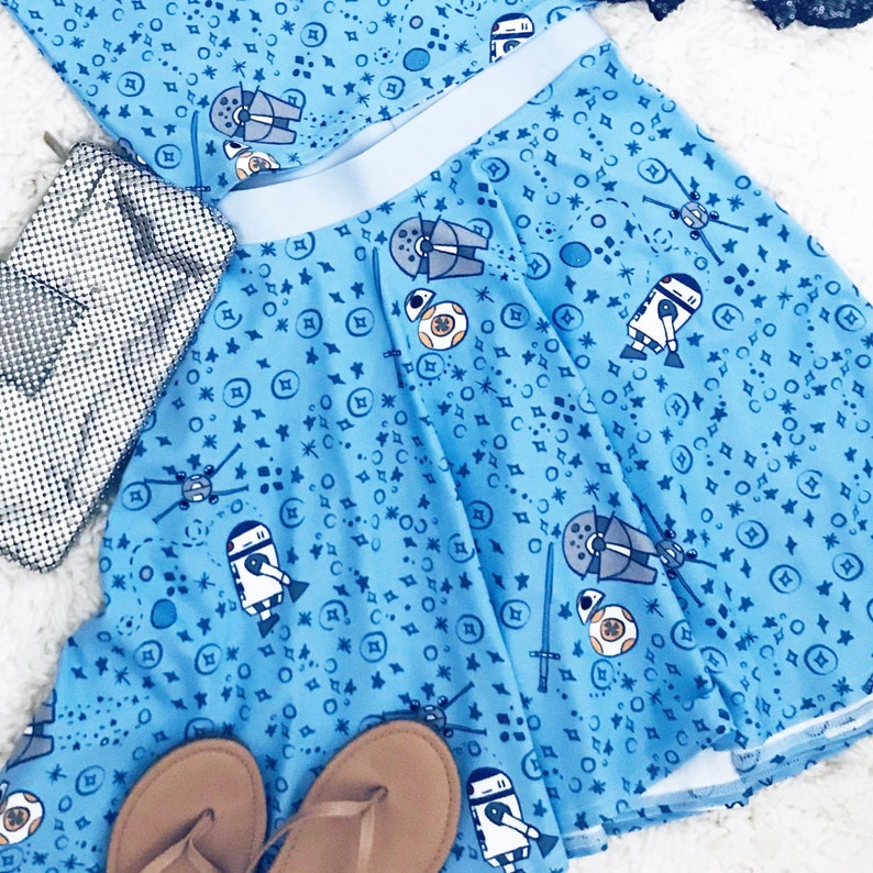 Puede incluir: Una falda azul con un estampado blanco y azul de personajes de Star Wars. La falda tiene una cintura azul claro y un forro blanco. La falda se muestra con un bolso de mano de malla plateada y sandalias marrones.