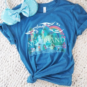 Puede incluir: Camiseta azul con un gráfico de Nunca Jamás, un arcoíris y el texto "Welcome to Neverland". Se ven un par de orejas de ratón azules con lentejuelas. La camiseta está anudada en la parte inferior.