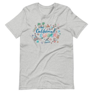May include: A heather grey t-shirt with a colorful doodle design featuring California Dreamin' text and various California-themed icons, including palm trees, a Ferris wheel, a castle, and a surfboard.