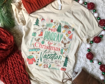 May include: A cream-coloured t-shirt featuring a Christmas graphic with the words "Hip Hooray for Christmas Vacation" and festive illustrations. A red knitted hat and scarf are visible, along with brown boots.